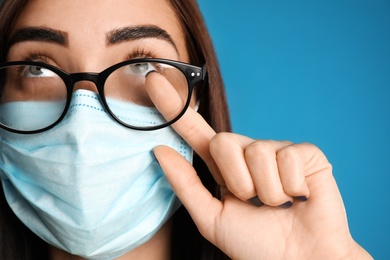 Young woman wiping foggy glasses caused by wearing disposable mask on blue background, closeup. Protective measure during coronavirus pandemic
