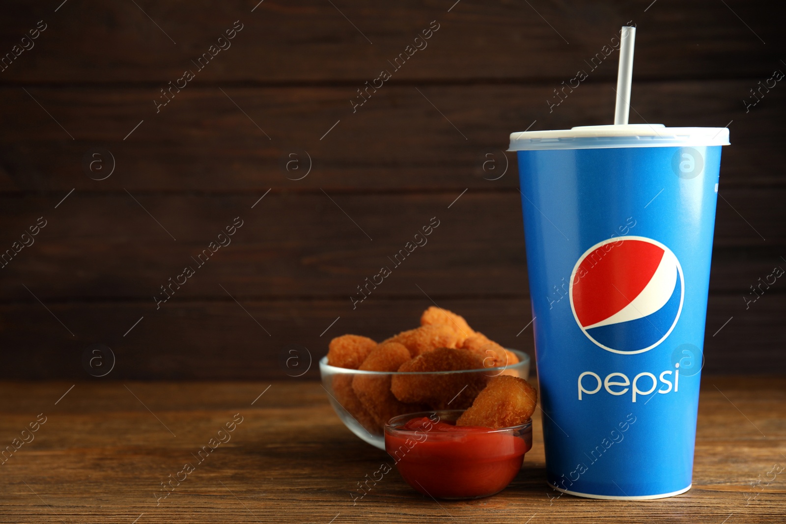 Photo of MYKOLAIV, UKRAINE - FEBRUARY 16, 2021: Cup of Pepsi, chicken nuggets and dip sauce on wooden table. Space for text