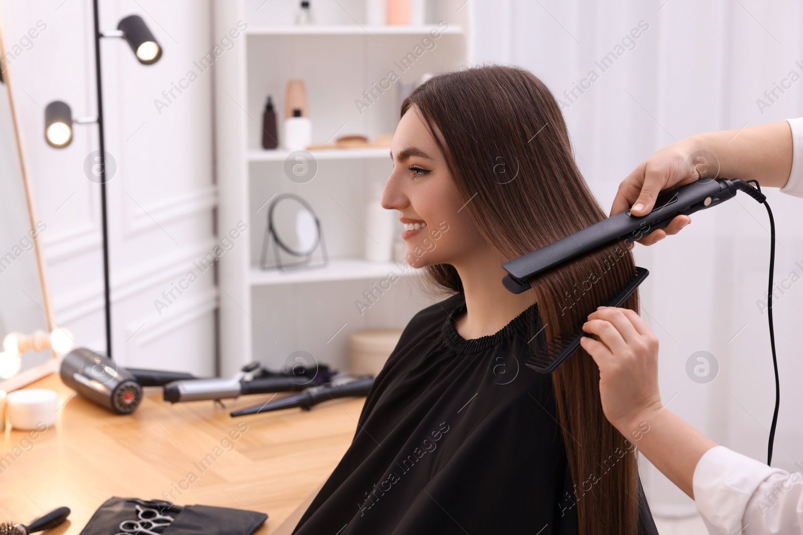 Photo of Hairdresser straightening woman's hair with flat iron in salon, closeup