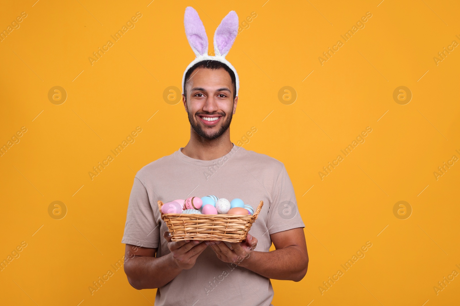 Photo of Happy African American man in bunny ears headband holding wicker tray with Easter eggs on orange background