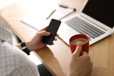 Man with cup of coffee and smartphone at workplace, closeup