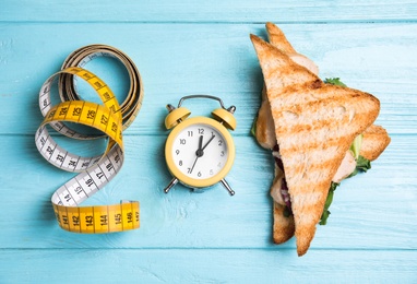 Tasty sandwich, alarm clock and measuring tape on light blue wooden table, flat lay. Diet regime