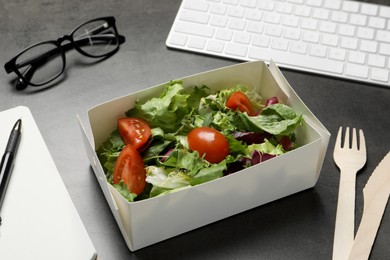 Photo of Container of tasty food, keyboard, glasses, cutlery and notebook on grey table. Business lunch