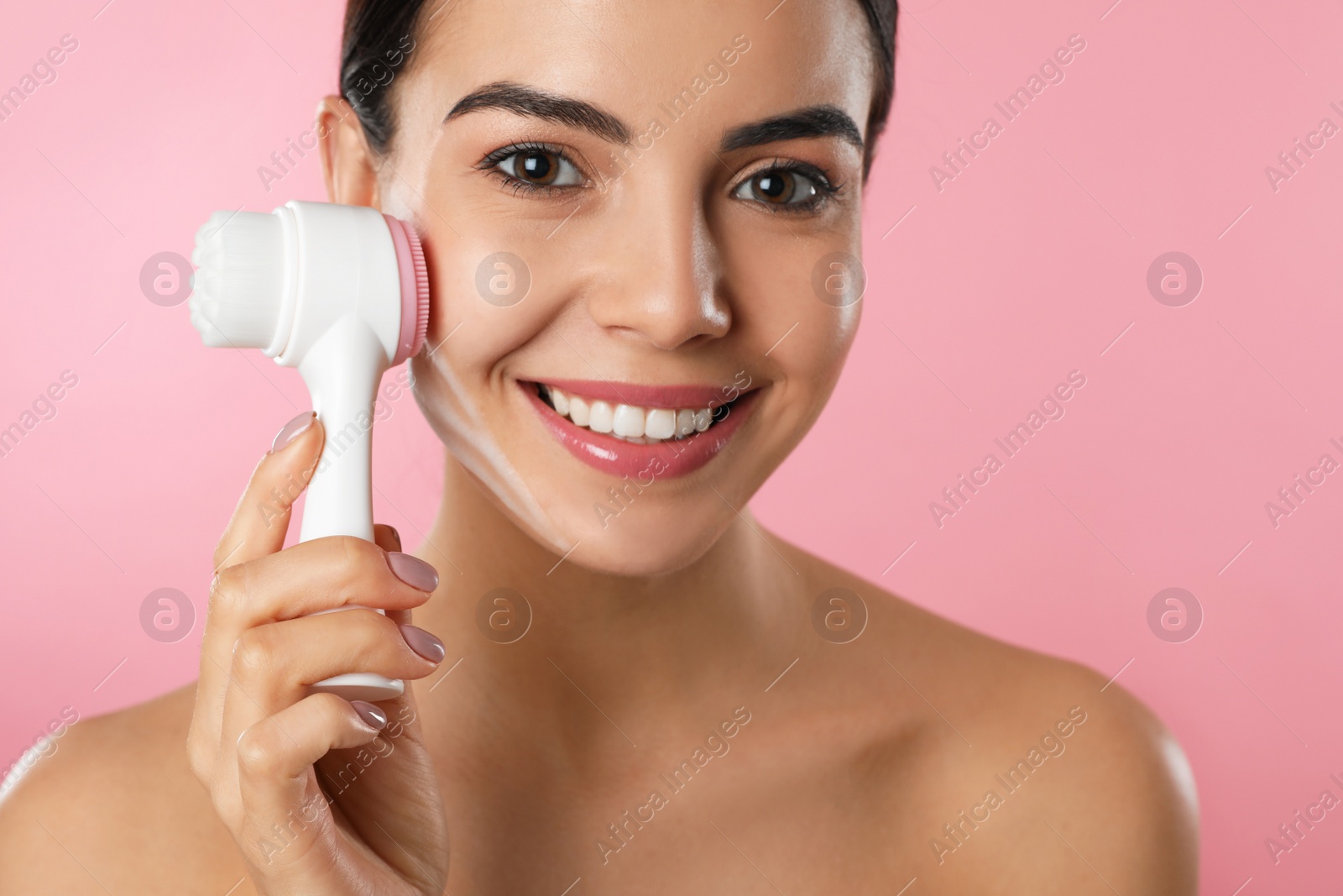 Photo of Young woman using facial cleansing brush on pink background, closeup. Washing accessory