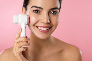 Photo of Young woman using facial cleansing brush on pink background, closeup. Washing accessory