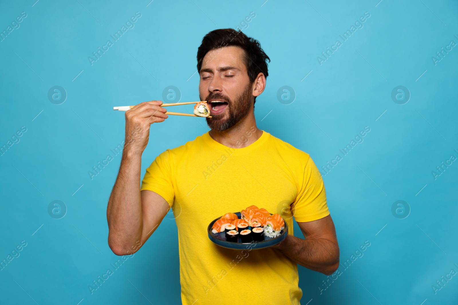 Photo of Man eating tasty sushi roll with chopsticks on light blue background