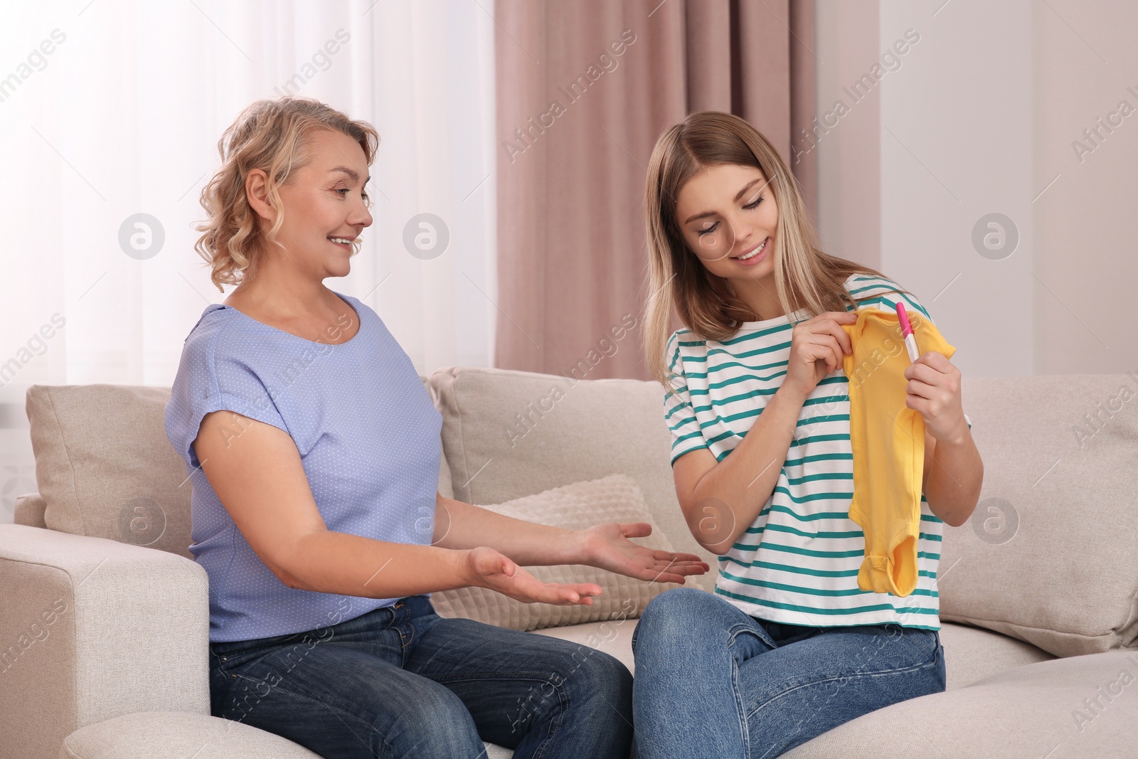 Photo of Young woman showing her mother baby's bodysuit and pregnancy test at home. Grandparent reaction to future grandson