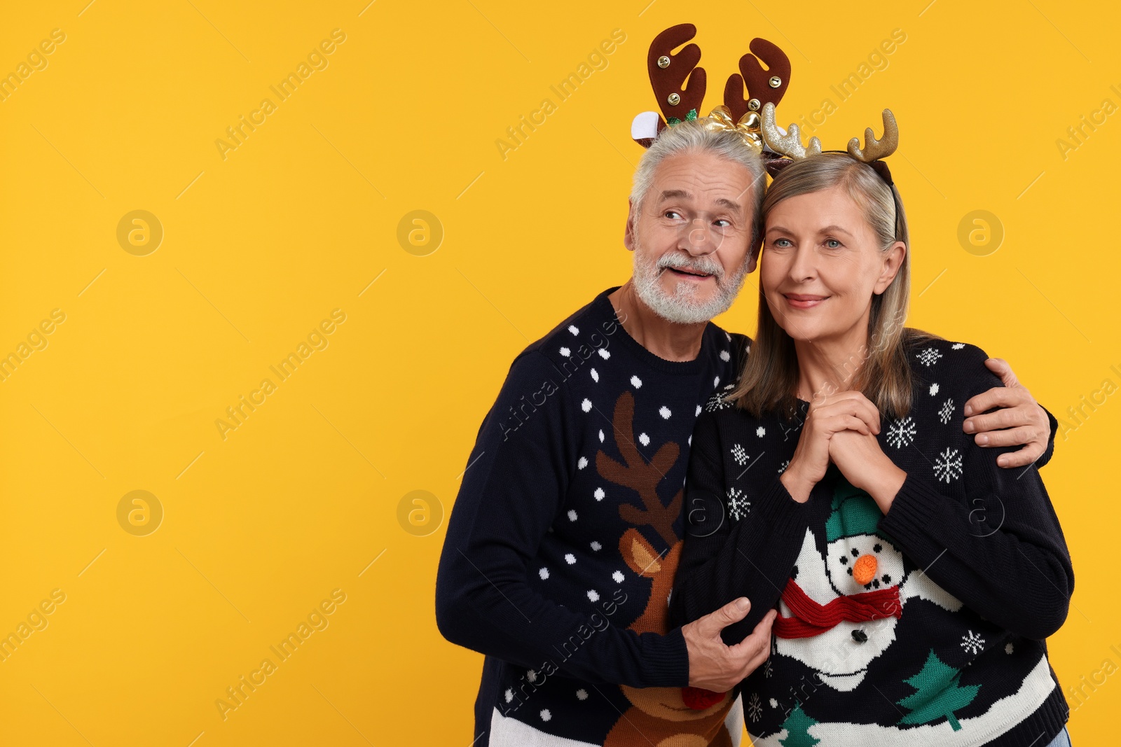 Photo of Senior couple in Christmas sweaters and reindeer headbands on orange background. Space for text