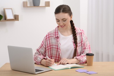 Photo of Happy young woman writing in notebook while working on laptop at wooden table indoors