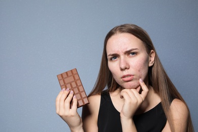 Young woman with acne problem holding chocolate bar on color background. Skin allergy