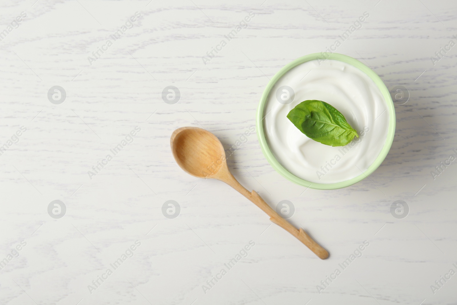 Photo of Bowl of fresh sour cream with basil and spoon on white wooden table, flat lay. Space for text