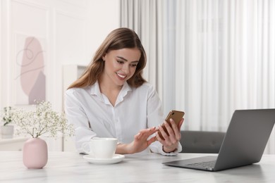 Happy woman with smartphone and laptop at white table in room