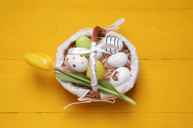 Wicker basket with festively decorated Easter eggs and beautiful tulip on yellow wooden background, top view