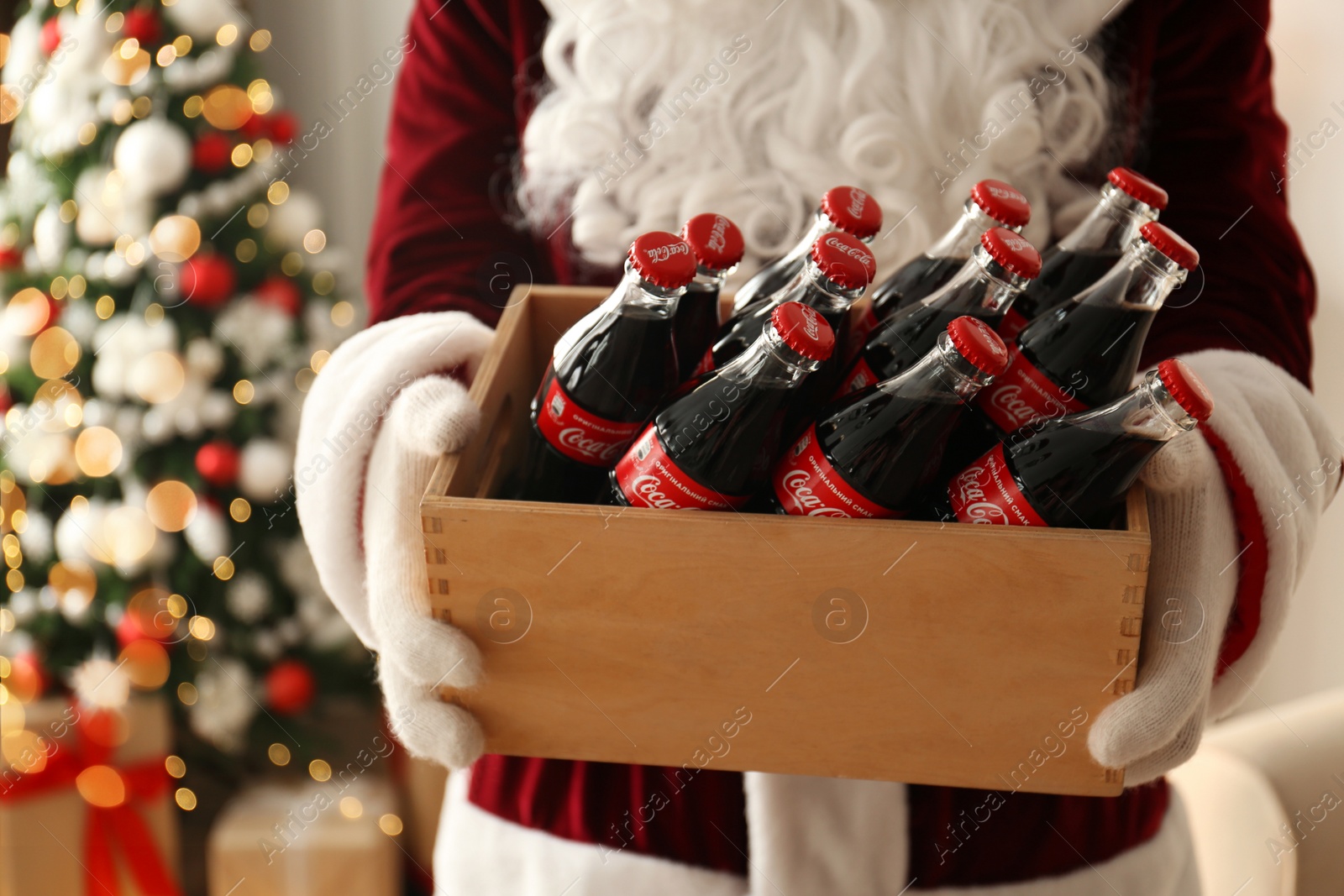 Photo of MYKOLAIV, UKRAINE - JANUARY 18, 2021: Santa Claus holding wooden crate full of Coca-Cola bottles in room with Christmas tree, closeup