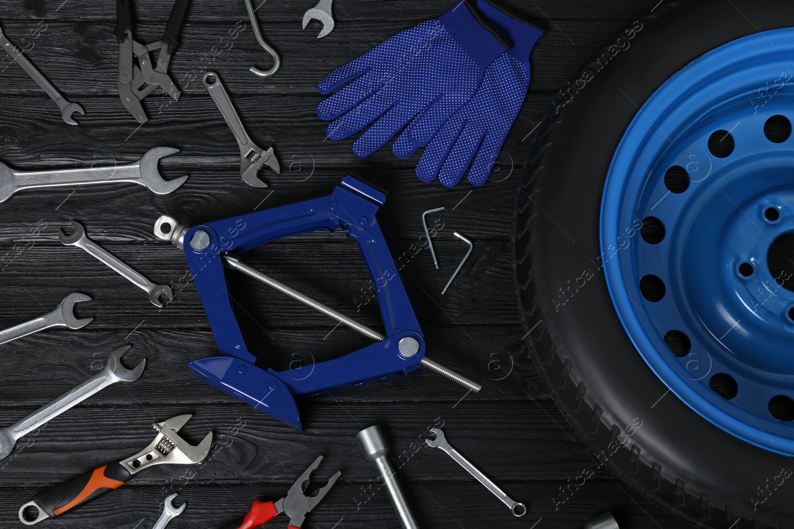 Photo of Car wheel, scissor jack, gloves and different tools on black wooden surface, flat lay