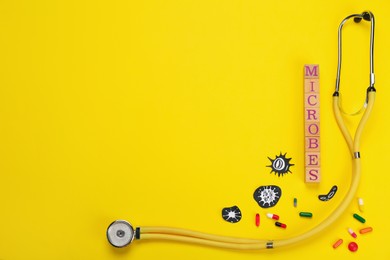 Word Microbes made with wooden cubes, colorful pills, pictures and stethoscope on yellow background, flat lay. Space for text