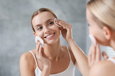 Photo of Smiling woman removing makeup with cotton pads in front of mirror indoors