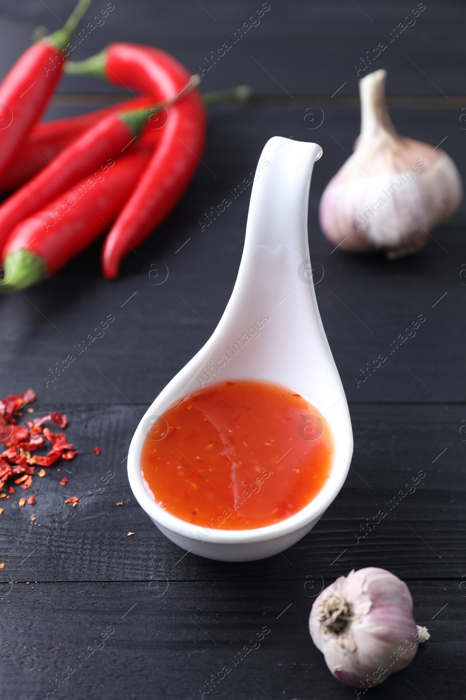 Photo of Spicy chili sauce in spoon, peppers and garlic on black wooden table, closeup