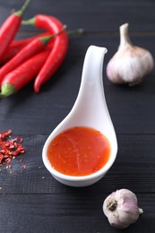 Spicy chili sauce in spoon, peppers and garlic on black wooden table, closeup