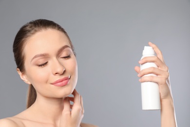 Photo of Young woman applying thermal water on face against grey background. Cosmetic product