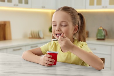 Photo of Cute little girl enjoying tasty yogurt at white marble table in kitchen