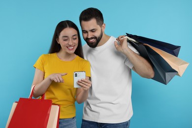 Happy couple with shopping bags and smartphone on light blue background