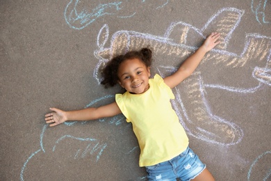 Photo of Little African-American child lying near chalk drawing of airplane on asphalt, top view