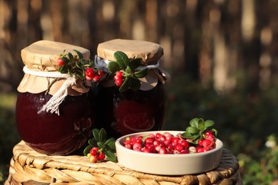 Photo of Jars of delicious lingonberry jam and red berries on wicker basket outdoors