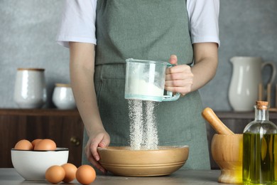 Photo of Woman sieving flour into bowl at table in kitchen, closeup