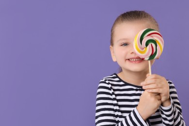Photo of Happy little girl covering eye with colorful lollipop swirl on violet background, space for text