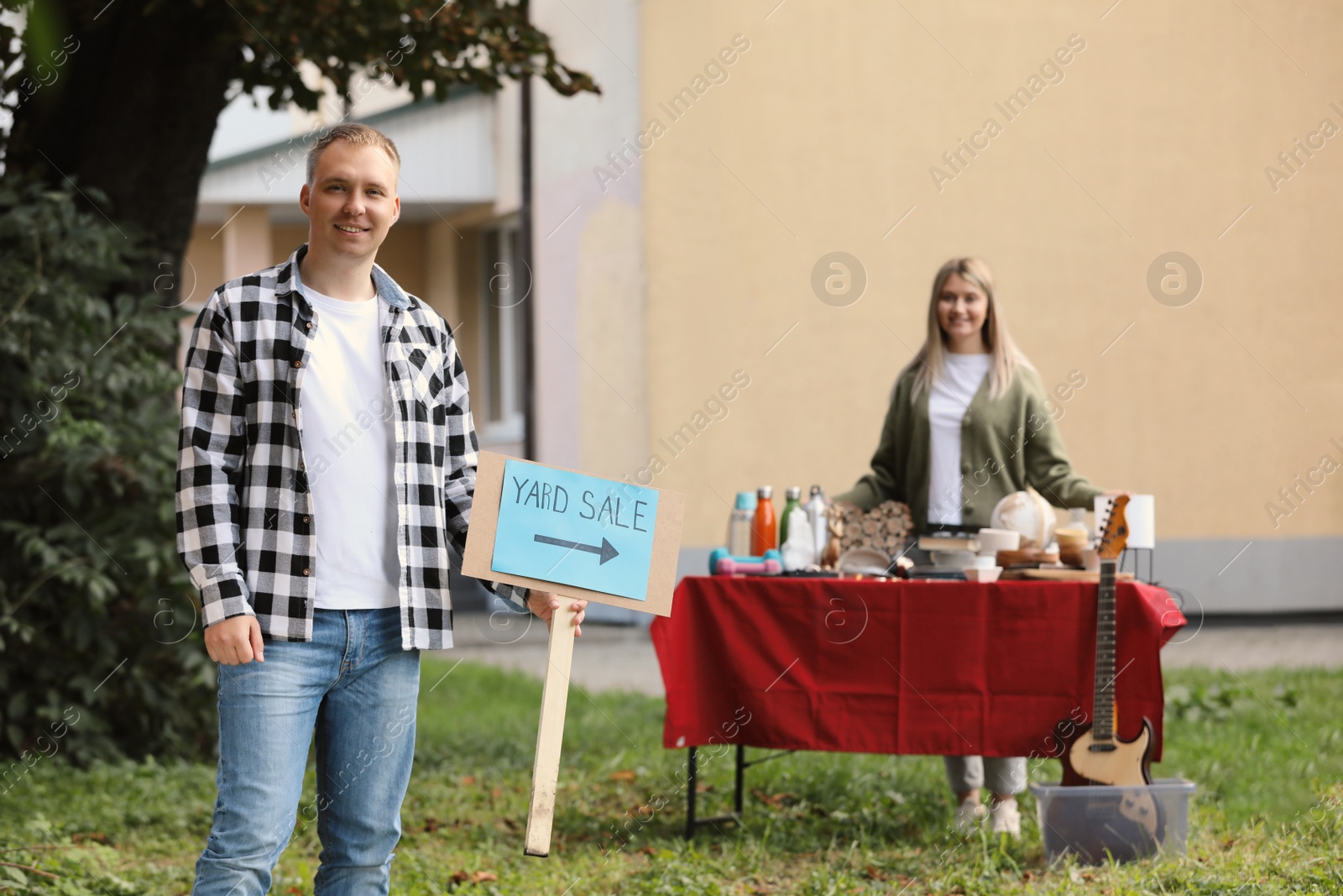 Photo of Man with sign Yard sale and woman near table of different items outdoors