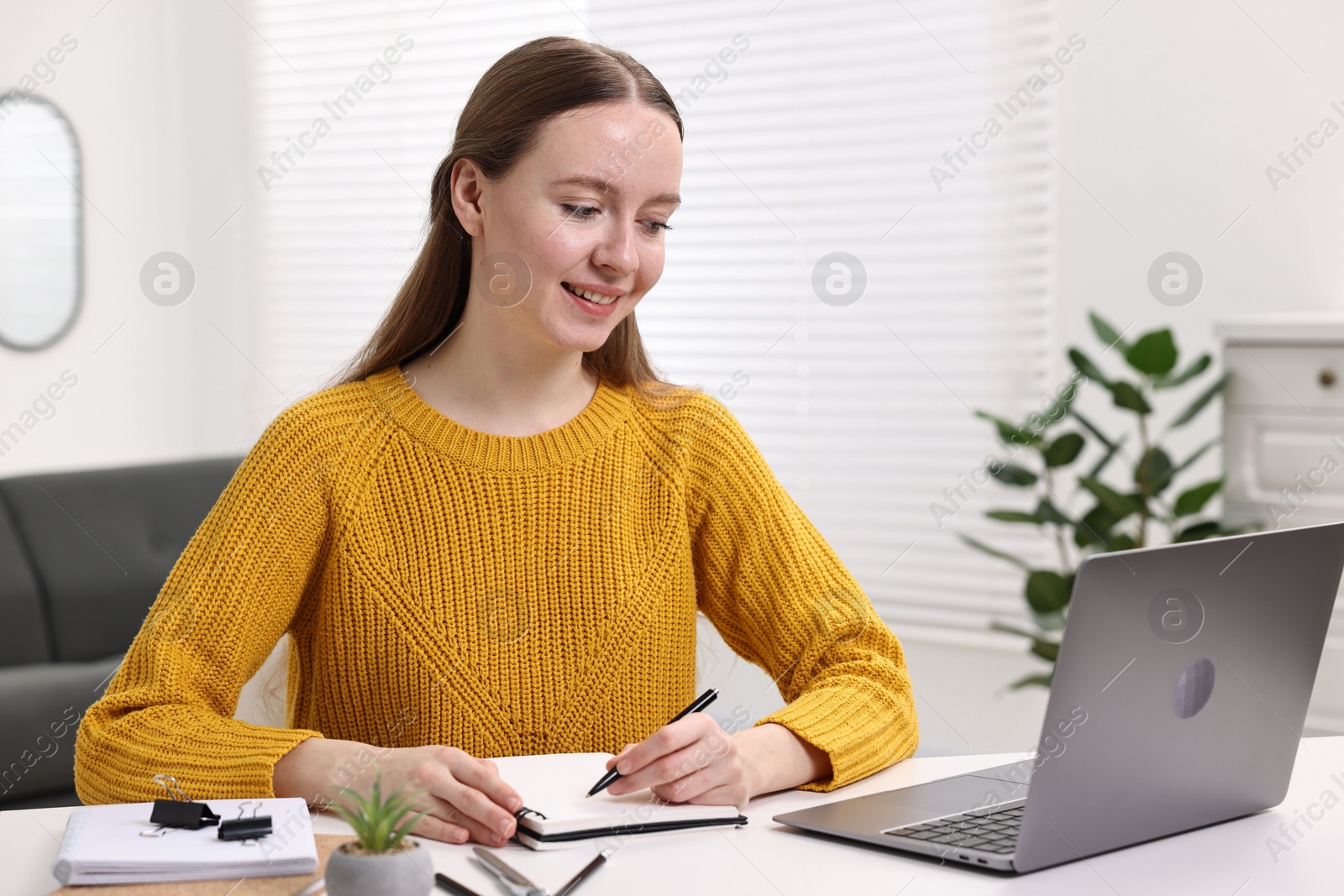 Photo of E-learning. Young woman taking notes during online lesson at white table indoors