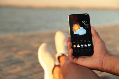 Man checking weather using app on smartphone near river, closeup. Data, sun with cloud and other illustrations on screen