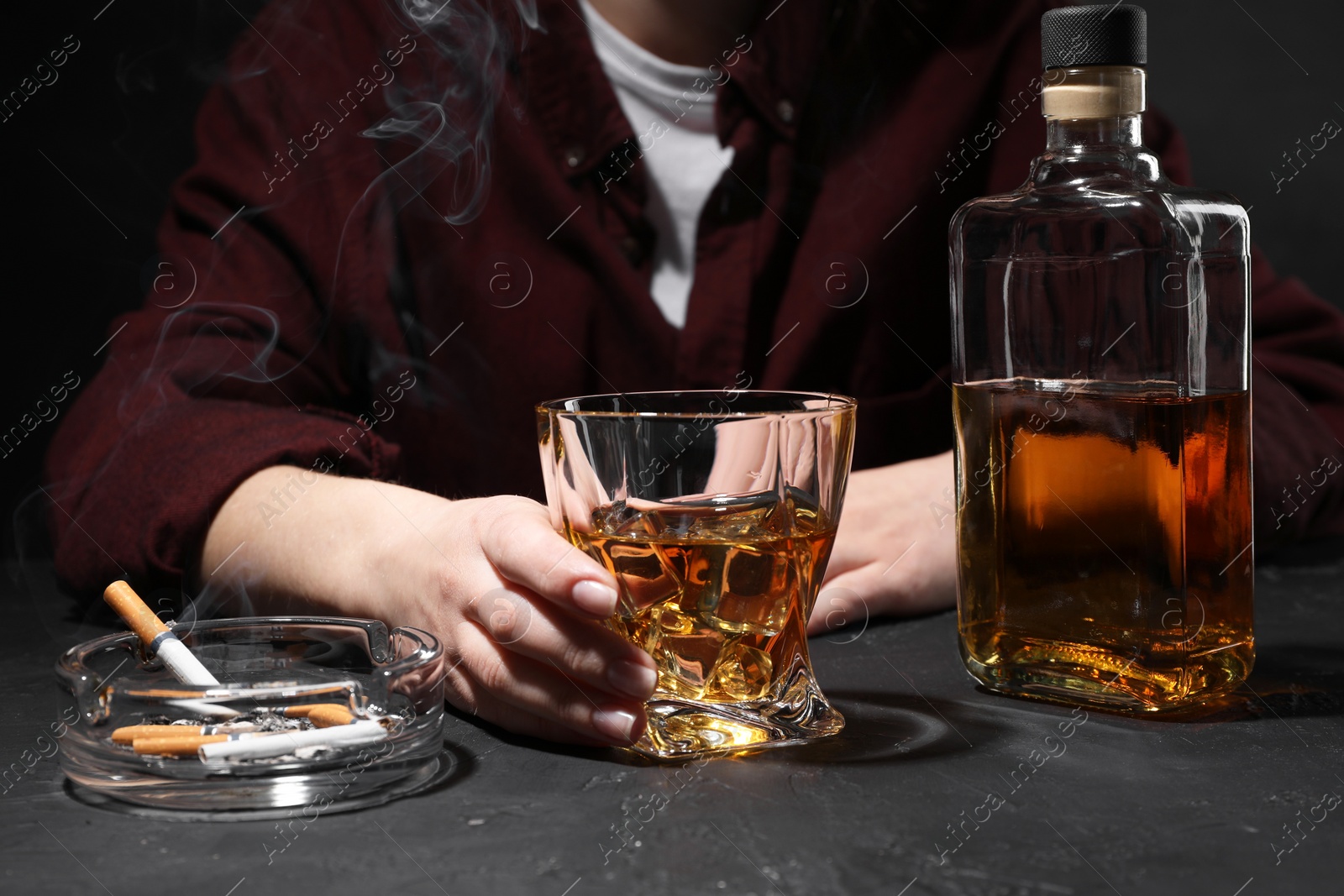 Photo of Alcohol addiction. Woman with whiskey and smoldering cigarettes at dark textured table, closeup