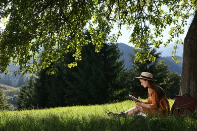 Young woman reading book under tree on meadow in mountains