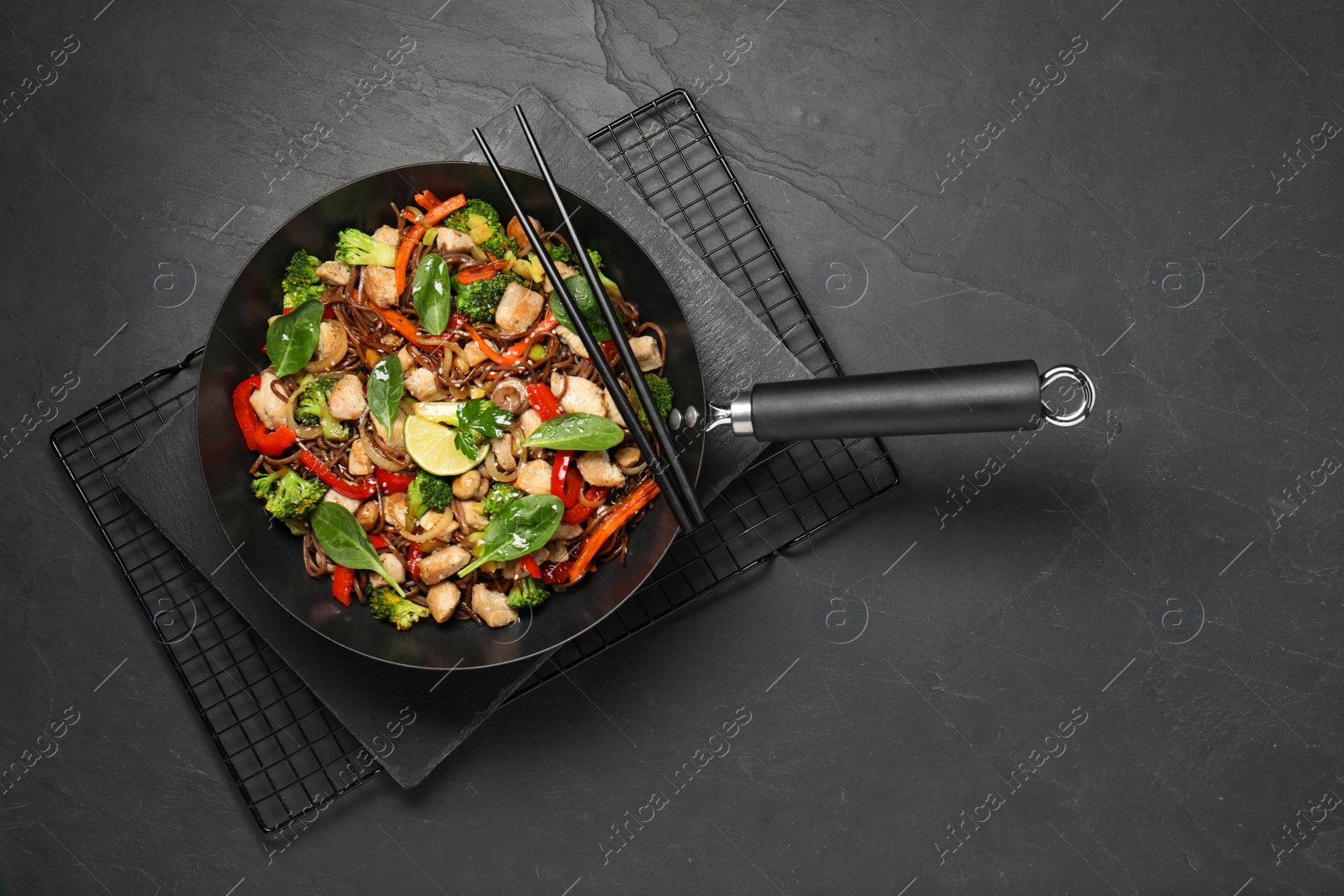 Photo of Stir-fry. Tasty noodles with meat in wok and chopsticks on grey textured table, top view