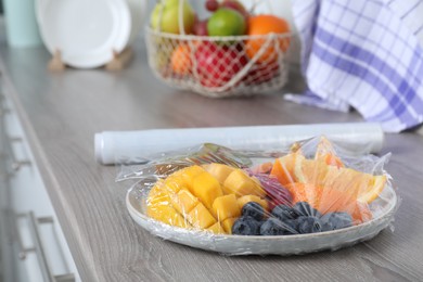 Photo of Plate of fresh fruits and berries with plastic food wrap on wooden table indoors