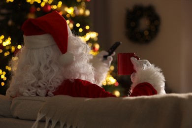 Photo of Merry Christmas. Santa Claus with cupdrink changing TV channels on sofa at home