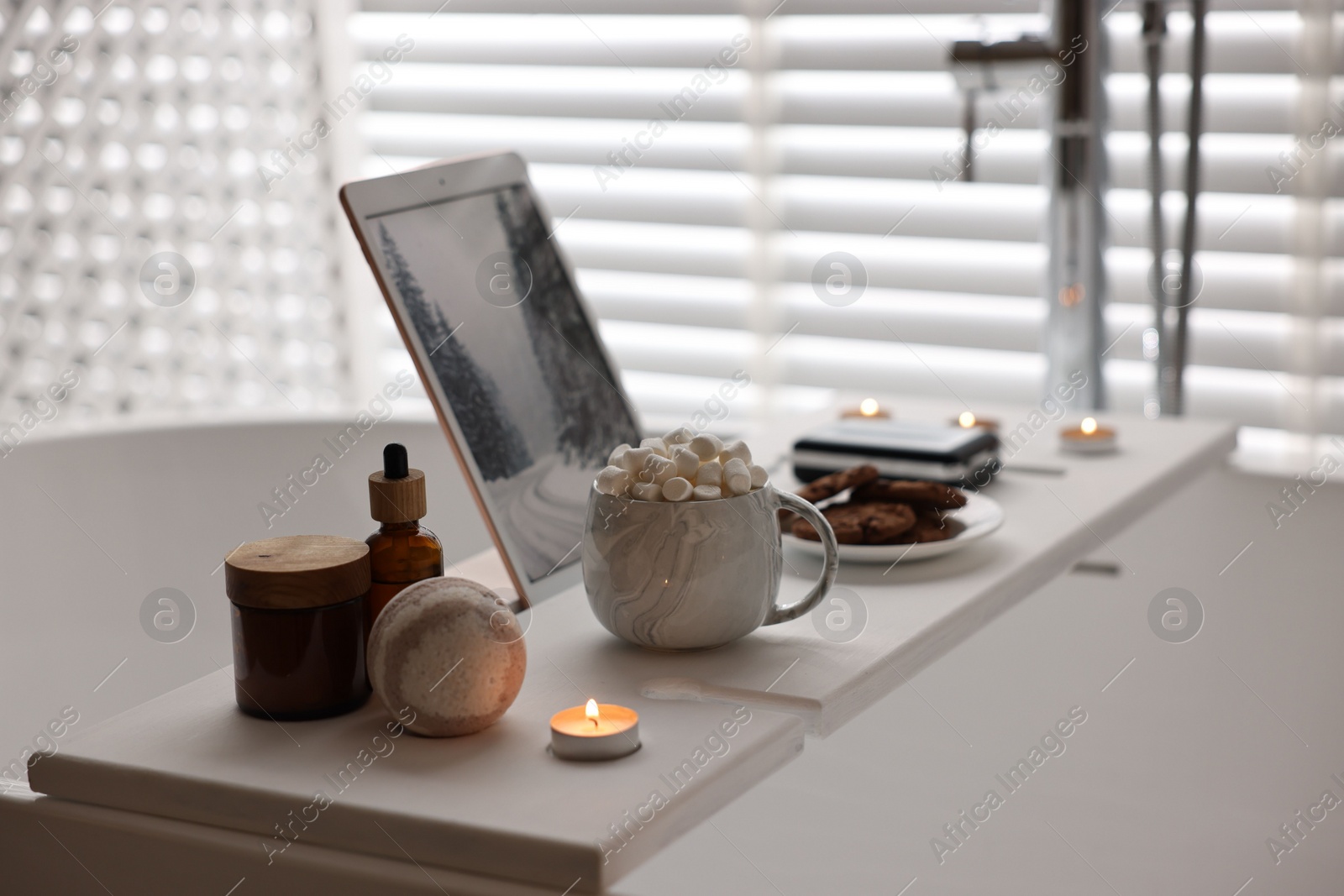 Photo of White wooden tray with tablet, spa products, hot drink and burning candle on bathtub in bathroom