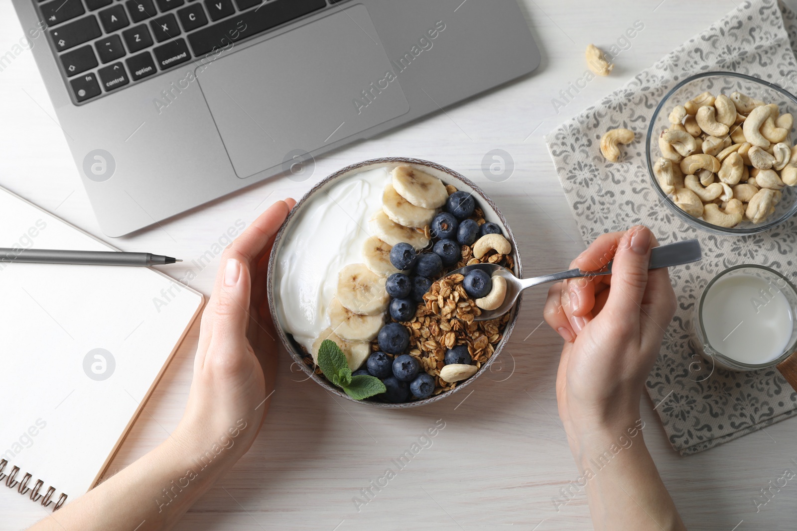 Photo of Woman eating tasty granola at white wooden table with laptop, top view