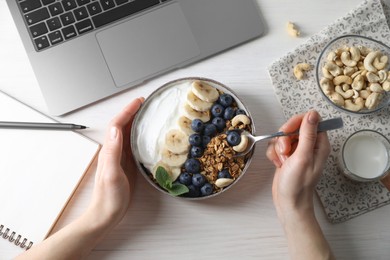 Photo of Woman eating tasty granola at white wooden table with laptop, top view