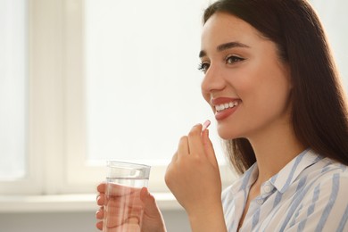 Photo of Young woman with glass of water taking dietary supplement pill indoors, space for text