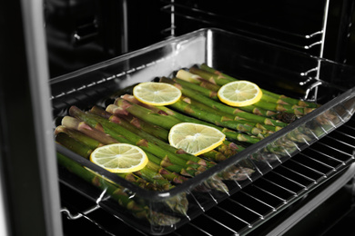 Photo of Raw asparagus with lemon slices in glass baking dish on oven rack, closeup