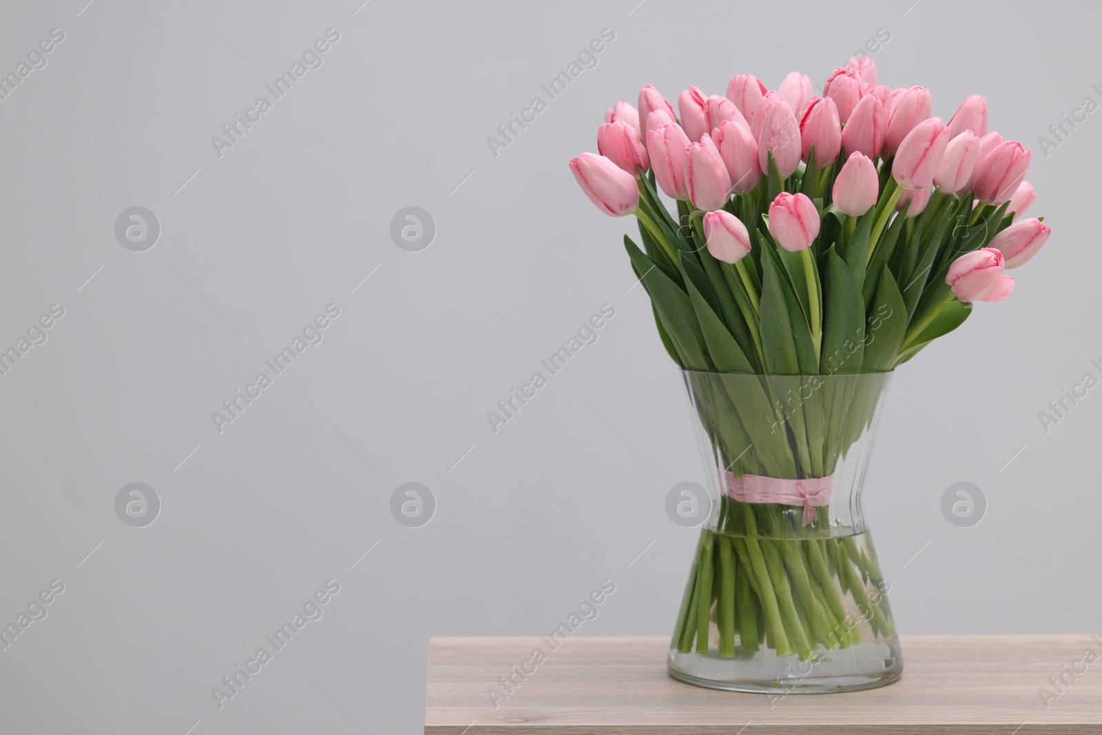 Photo of Bouquet of beautiful pink tulips in vase on wooden table against grey background, space for text