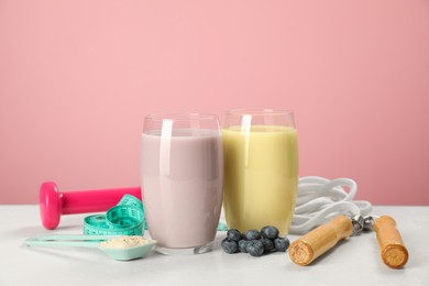 Photo of Tasty shakes with blueberries, sports equipment, measuring tape and powder on white table against pink background. Weight loss