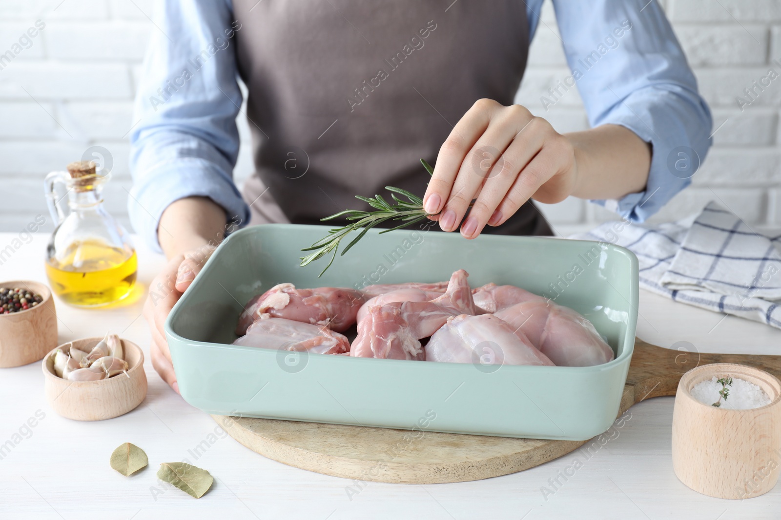 Photo of Woman putting rosemary into baking dish with raw rabbit meat at white wooden table, closeup