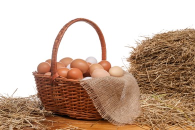 Photo of Wicker basket full of chicken eggs and dried straw on wooden table against white background