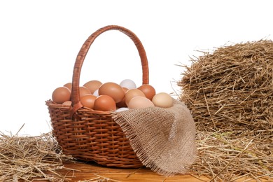 Wicker basket full of chicken eggs and dried straw on wooden table against white background