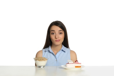 Photo of Doubtful woman choosing between yogurt with granola and cake at table on white background