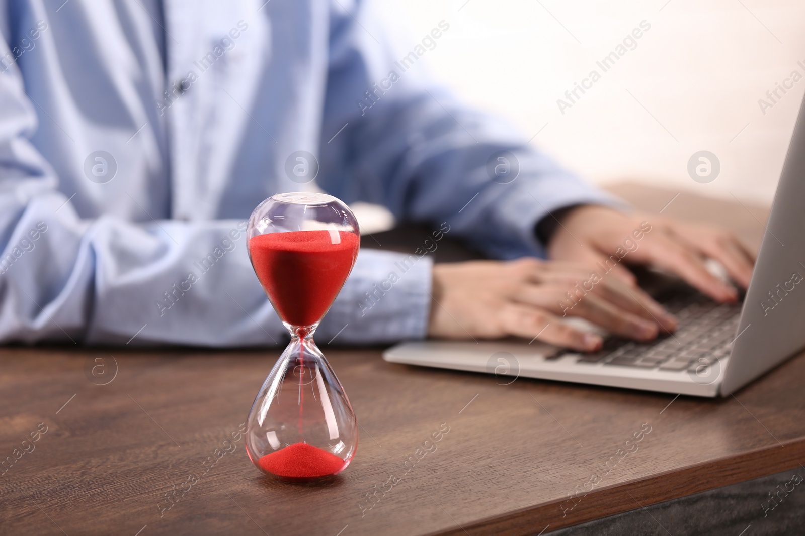 Photo of Hourglass with flowing sand on wooden table, selective focus. Man using laptop indoors